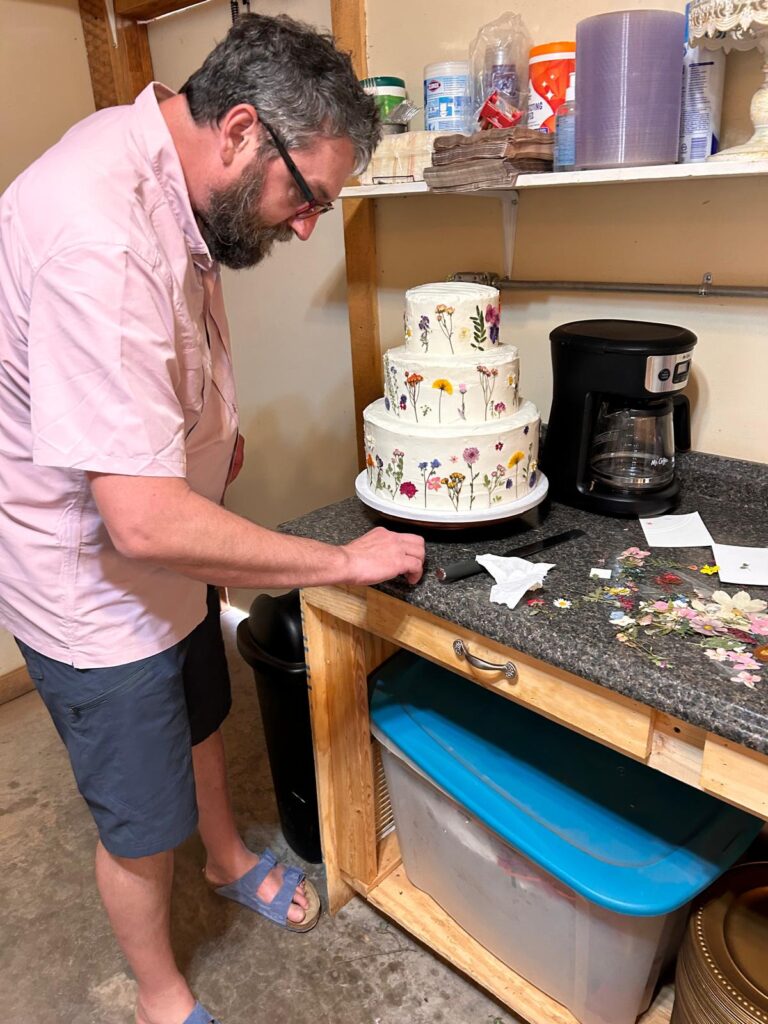 A man helping place dried flowers onto a white frosted three teir wedding cake.