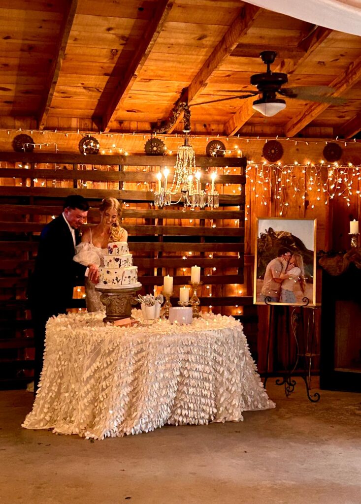 This image contains a three teir wedding cake with dried flowers on it in a barn wedding venue. The cake is being cut by the married couple.