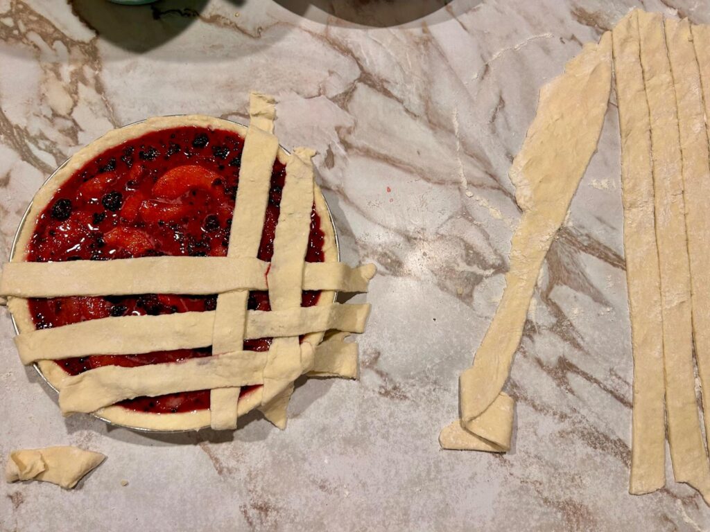 Pie plate lined with homemade pie crust, filled with peach and blackberry pie filling. The top pie crust is being weaved together.